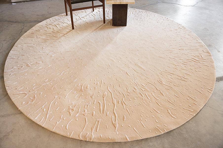A full view of a large, round beige carpet featuring subtle raised splatter designs, paired with a wooden chair and table, set in a contemporary, open space.
