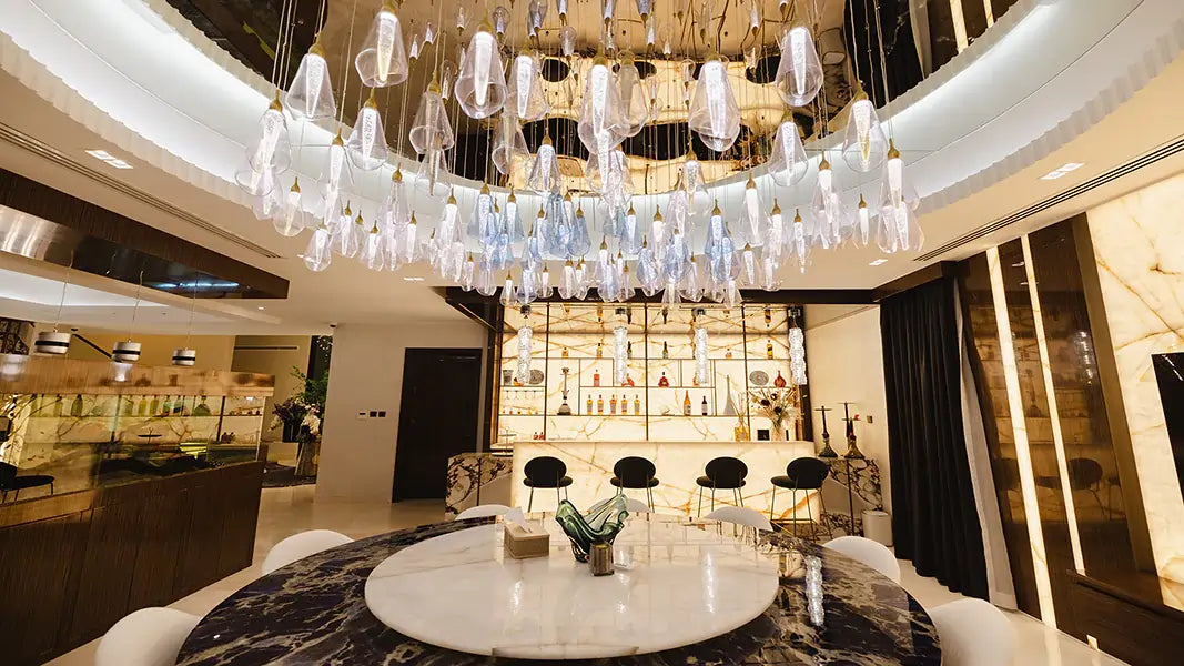 Luxurious modern dining room with a large circular table, marble flooring, and a stunning glass chandelier. The room features a wet bar with backlit shelves and stylish bar stools.