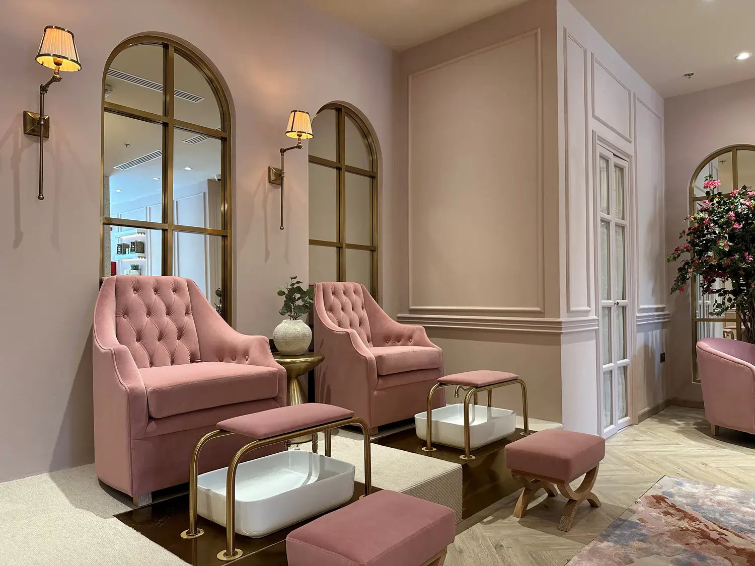 Luxurious spa pedicure chairs with pink tufted upholstery and gold accents, set against a soft blush wall with elegant wall-mounted lamps. The space exudes a chic, modern interior design with arched mirrors, accentuating a relaxing and stylish ambiance perfect for a high-end spa experience.