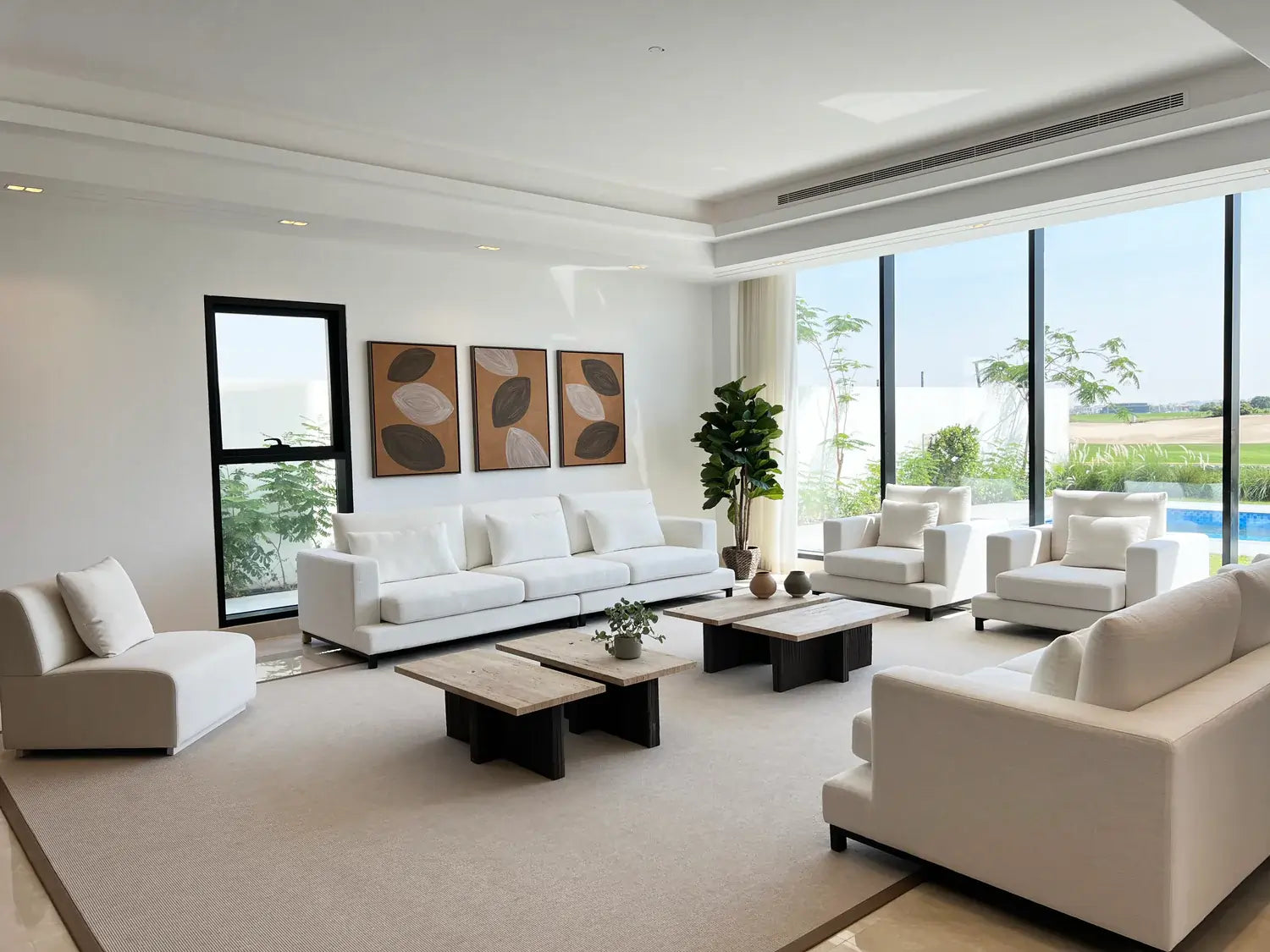 Modern living room with minimalist white sofas and armchairs, accented by abstract wall art and a large potted plant. Floor-to-ceiling windows provide natural light and offer views of a lush garden and pool, creating a serene and open atmosphere ideal for relaxation and contemporary interior design.